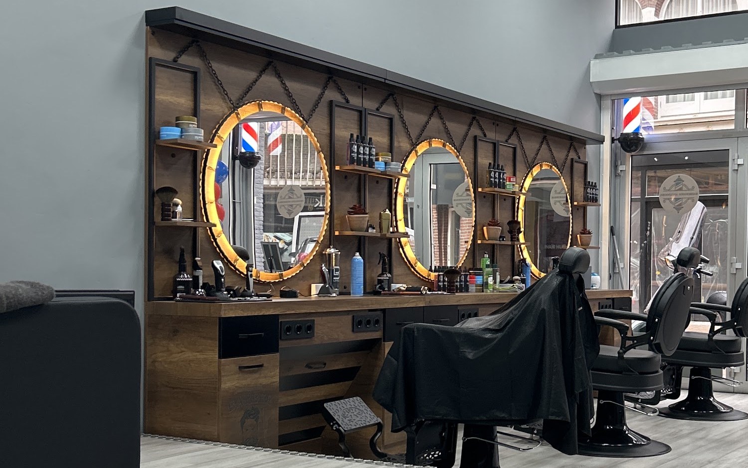 De houten spiegelwand met drie ronde, verlichte spiegels, planken met producten en de zwarte barberstoelen